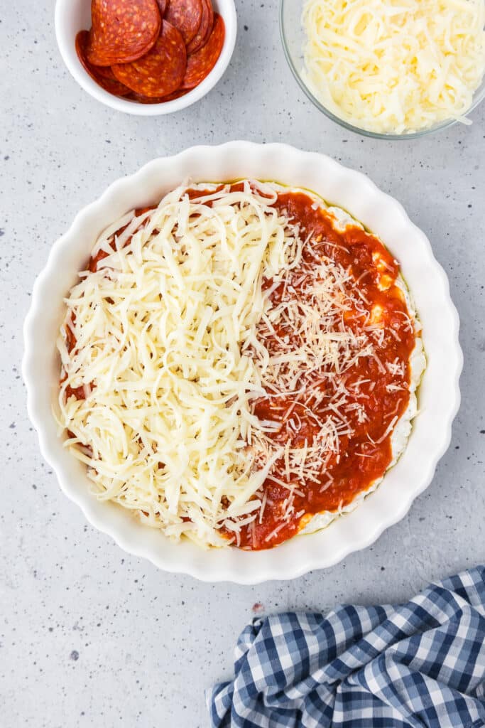 Pepperoni dip in a round dish with cheese being spread over marinara.