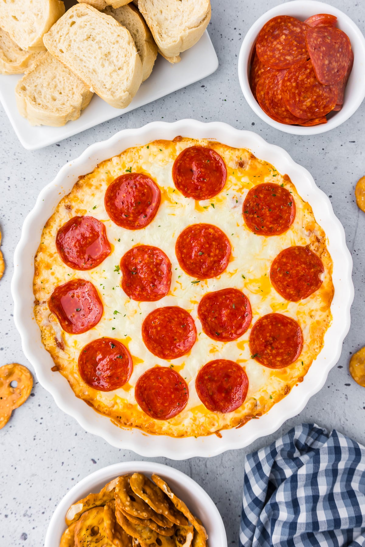 Pepperoni dip in a white round pie dish with pretzels and bread nearby on the counter.