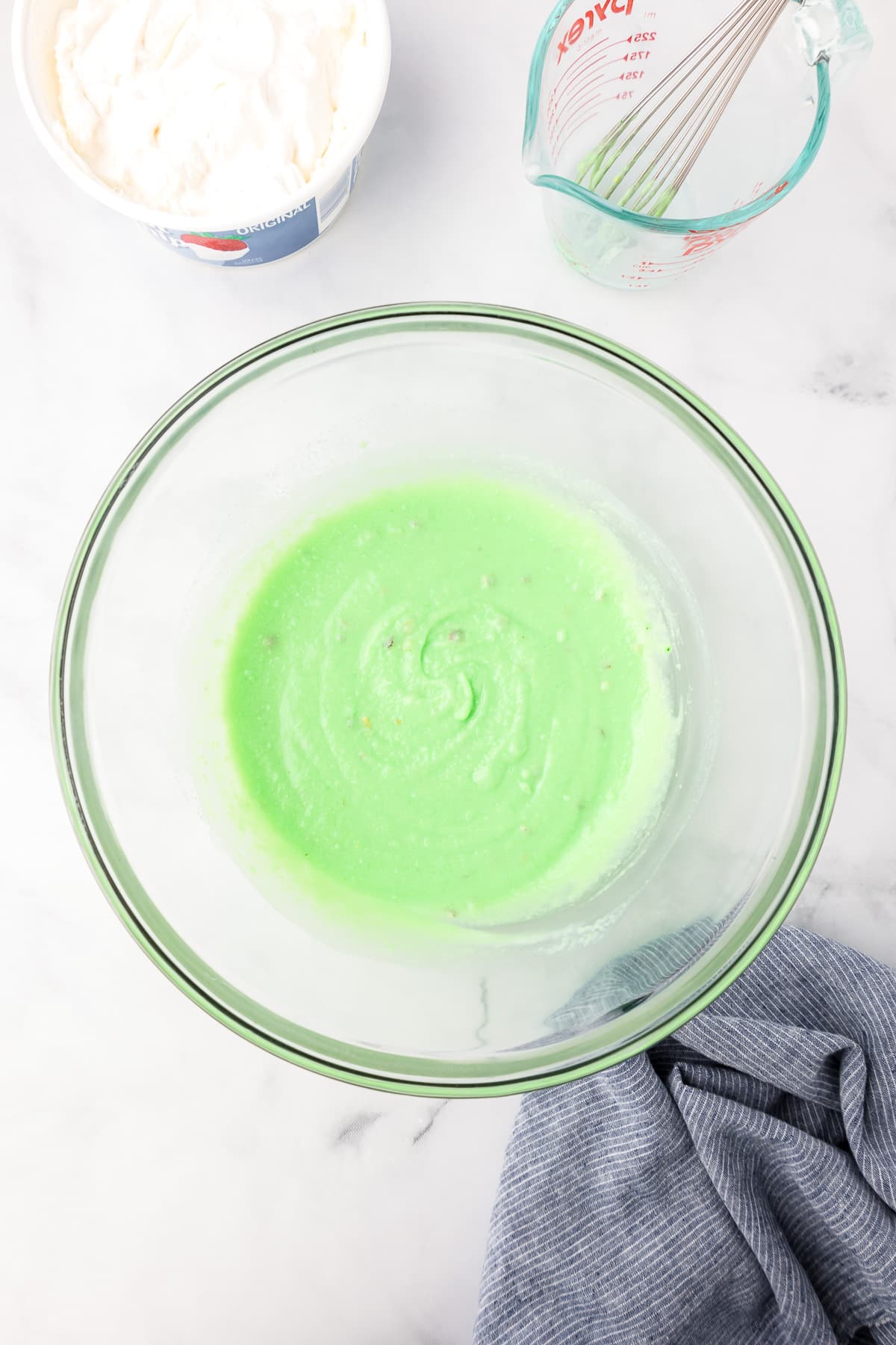 A bowl of green pistachio pudding next to a measuring cup with a whisk and a container of whipped topping.