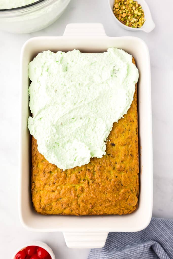 A white baking dish with a pistachio cake half covered in green frosting and a bowl of chopped pistachios nearby.