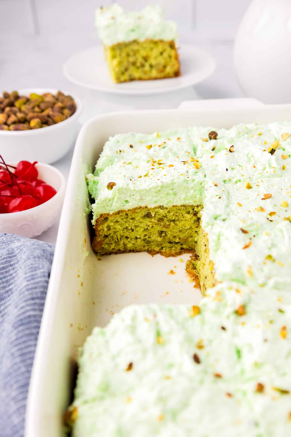 A white baking dish full of green frosted pistachio pudding cake with a slice of cake missing showing the cake underneath.
