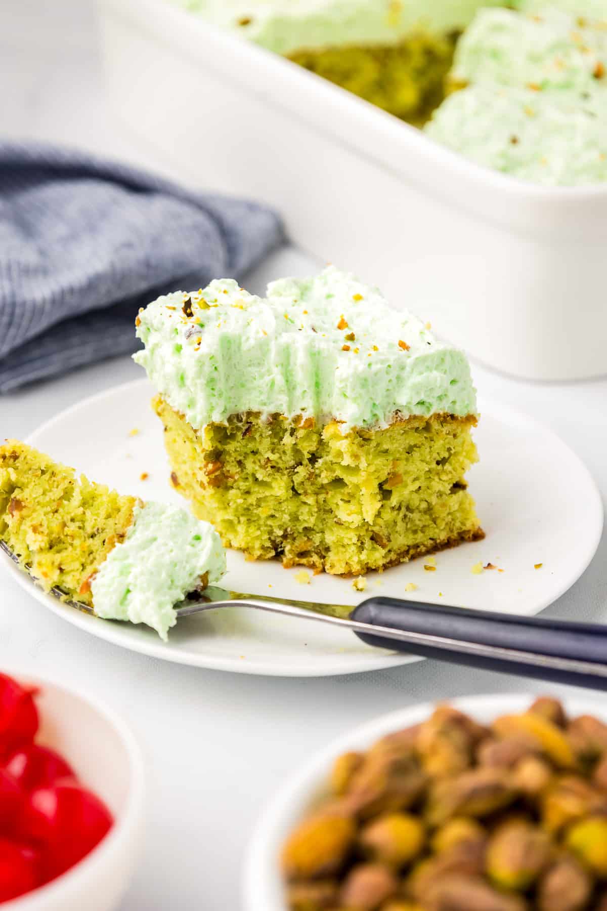 A slice of green pistachio cake topped with green frosting and chopped nuts on a plate.