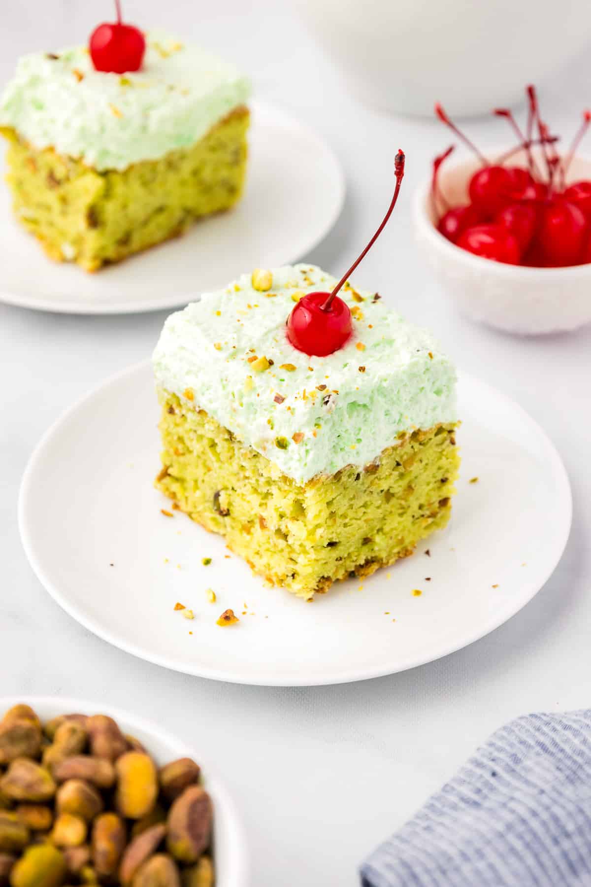 Pistachio pudding cake on a plate with a maraschino cherry and chopped pistachios on top.
