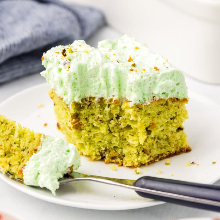 A slice of green frosted pistachio pudding cake on a plate with a bite removed and a fork on the plate with the bite.