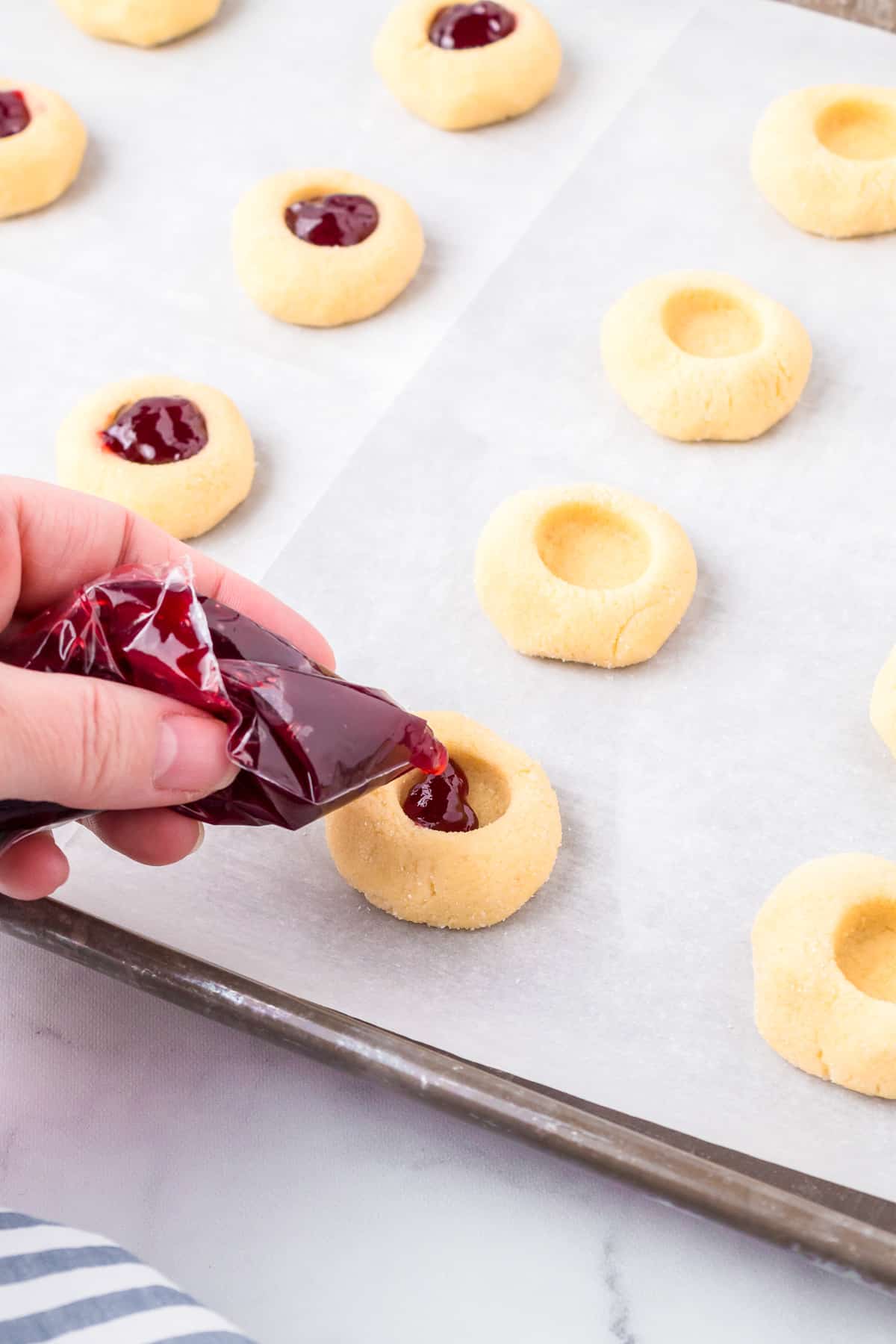 A person's hand filling the center of thumbprint cookies on a baking sheet with a bag filled with jam.