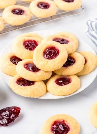 A plate of thumbprint cookies filled with raspberry jam with more cooling on a wire rack nearby.