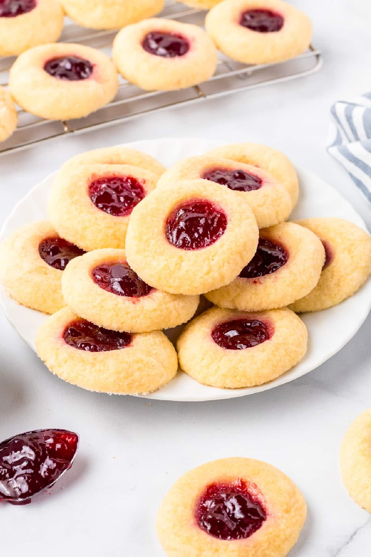 A plate of thumbprint cookies filled with raspberry jam with more cooling on a wire rack nearby.