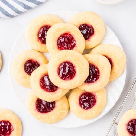 Raspberry thumbprint cookies on a plate from above with a wire cooling rack with more cookies nearby.