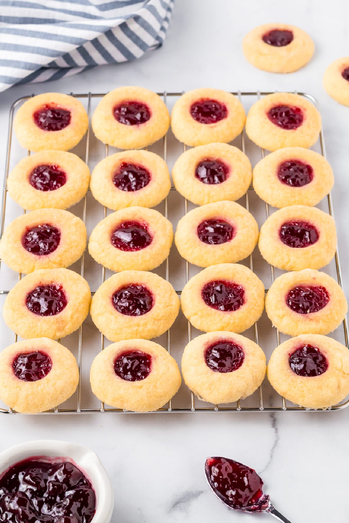 Raspberry jam thumbprint cookies in rows on a wire cooling rack with jam and a spoon dilled with jam on the counter nearby.