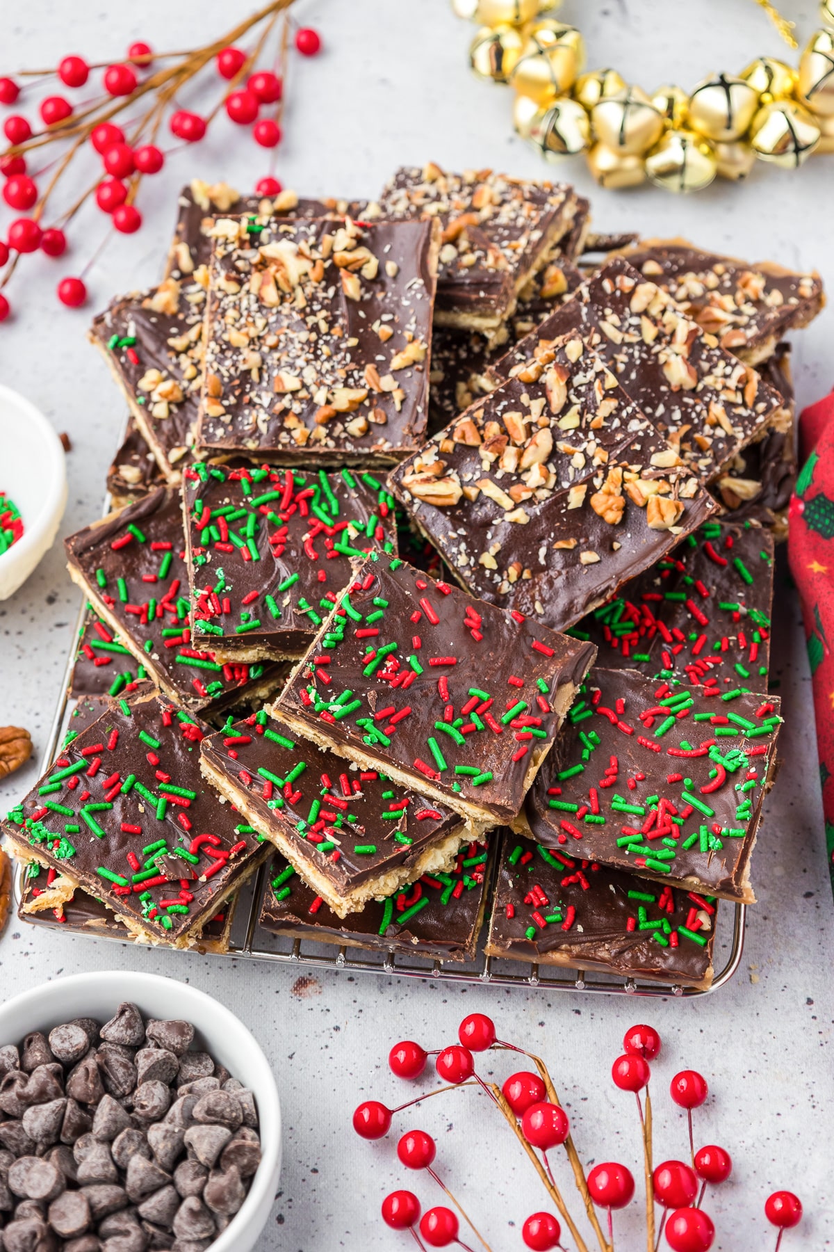 A tray of chocolate covered saltine toffee on a table, half covered in pecans half covered in red and green sprinkles.