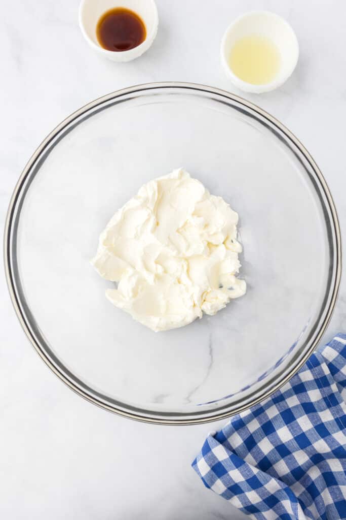 A bowl of cream cheese with small bowls of lemon juice and vanilla nearby on the counter.