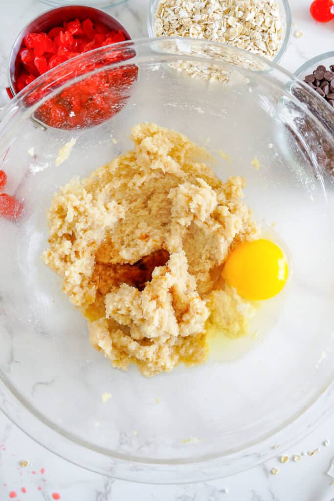 Creamed butter, sugar and egg being mixed together for a maraschino cherry cookie dough with chopped cherries, oats and chocolate chips in bowls nearby.