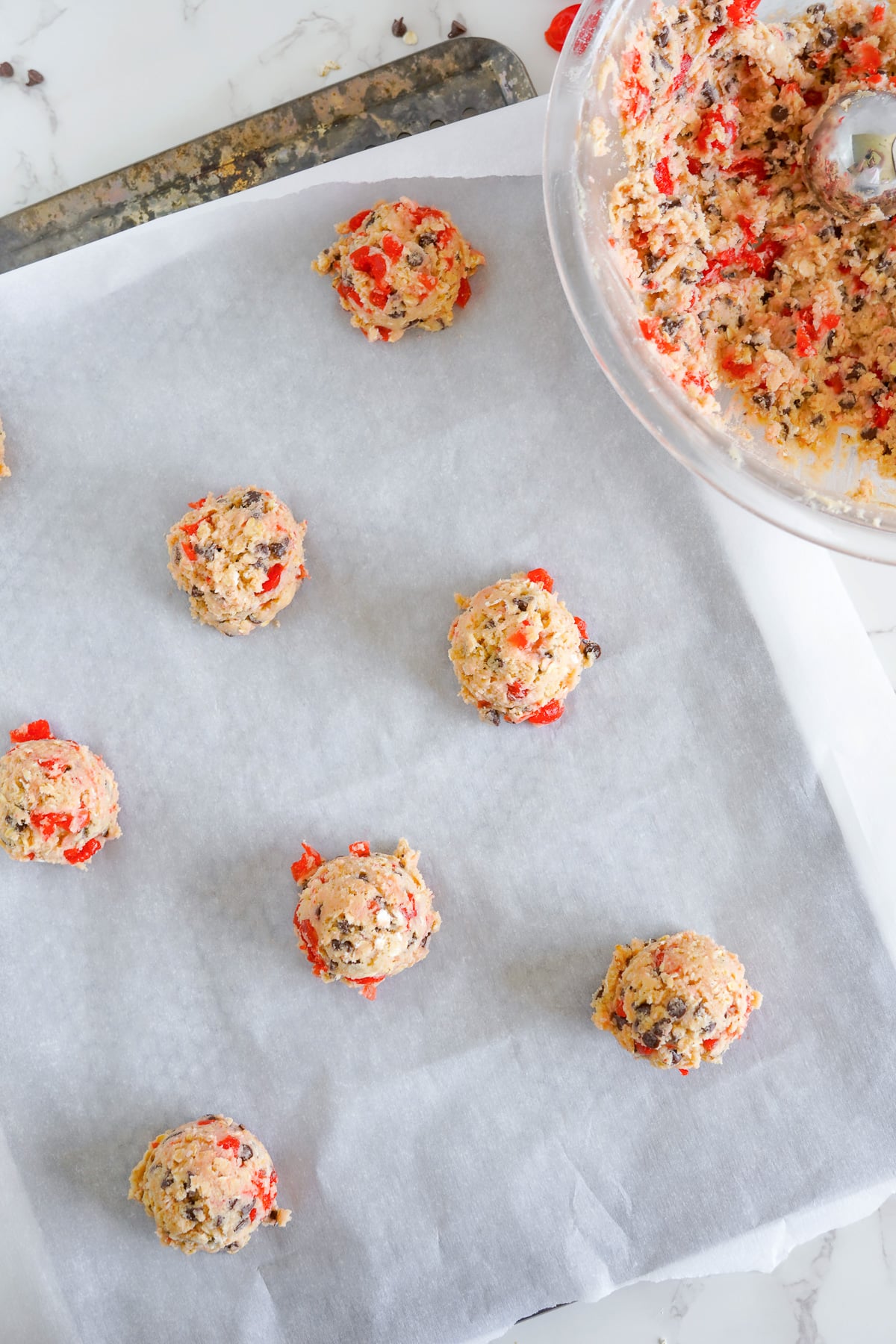 A baking sheet with maraschinio cherry cookies dough balls on it with a bowl of cookie dough nearby.