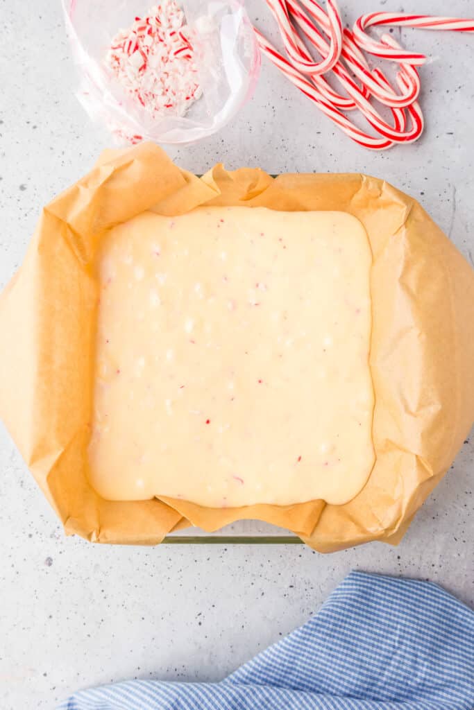 White chocolate peppermint fudge in a square baking pan lined with parchment paper with a bag of candy cane pieces and candy canes nearby on the counter.