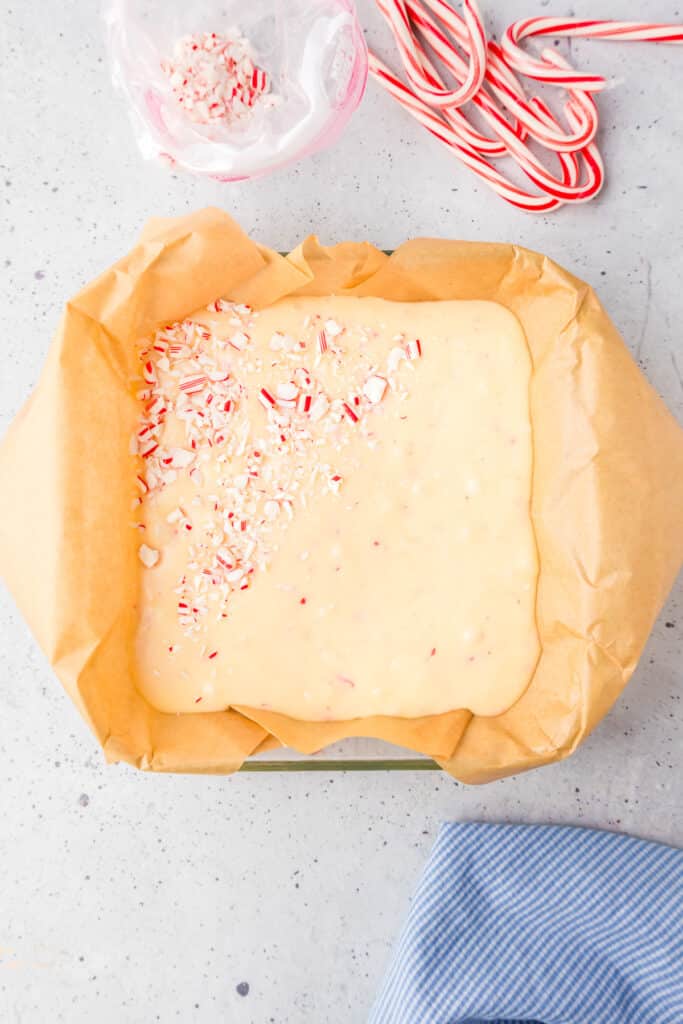 A square pan lined with parchment paper with liquid white fudge being sprinkled with candy cane bits.