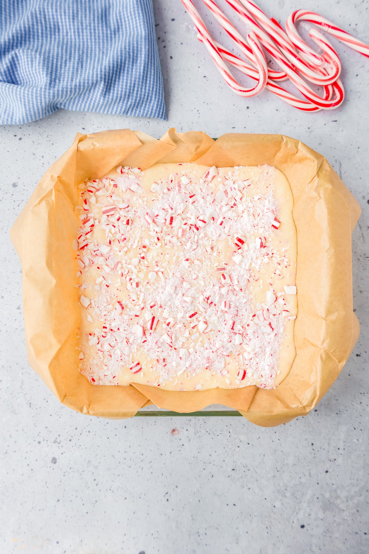 A square pan lined with parchment paper with white fudge after it is set topped with candy canes pieces on a counter.