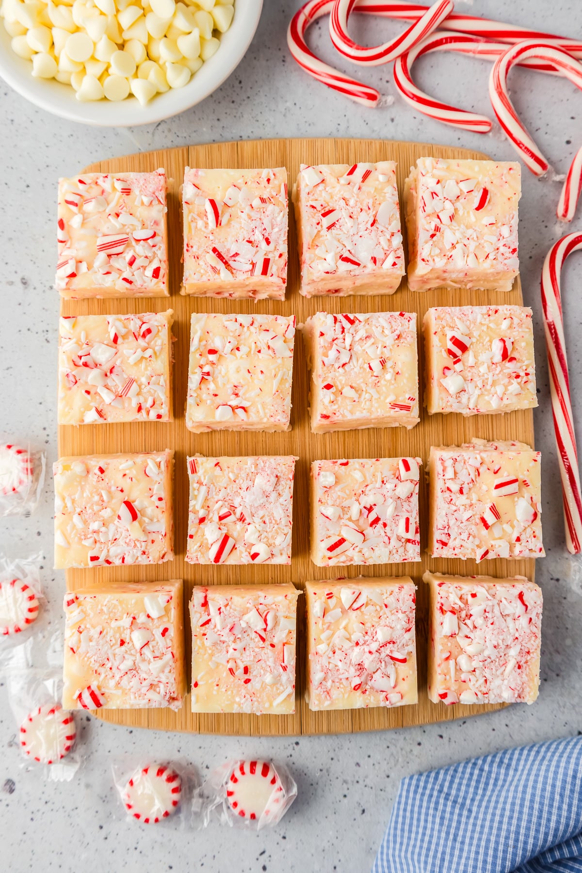 Candy cane fudge squares on a cutting board sliced into 16 pieces with more peppermints, white chocolate and candy canes next to the cutting board nearby..