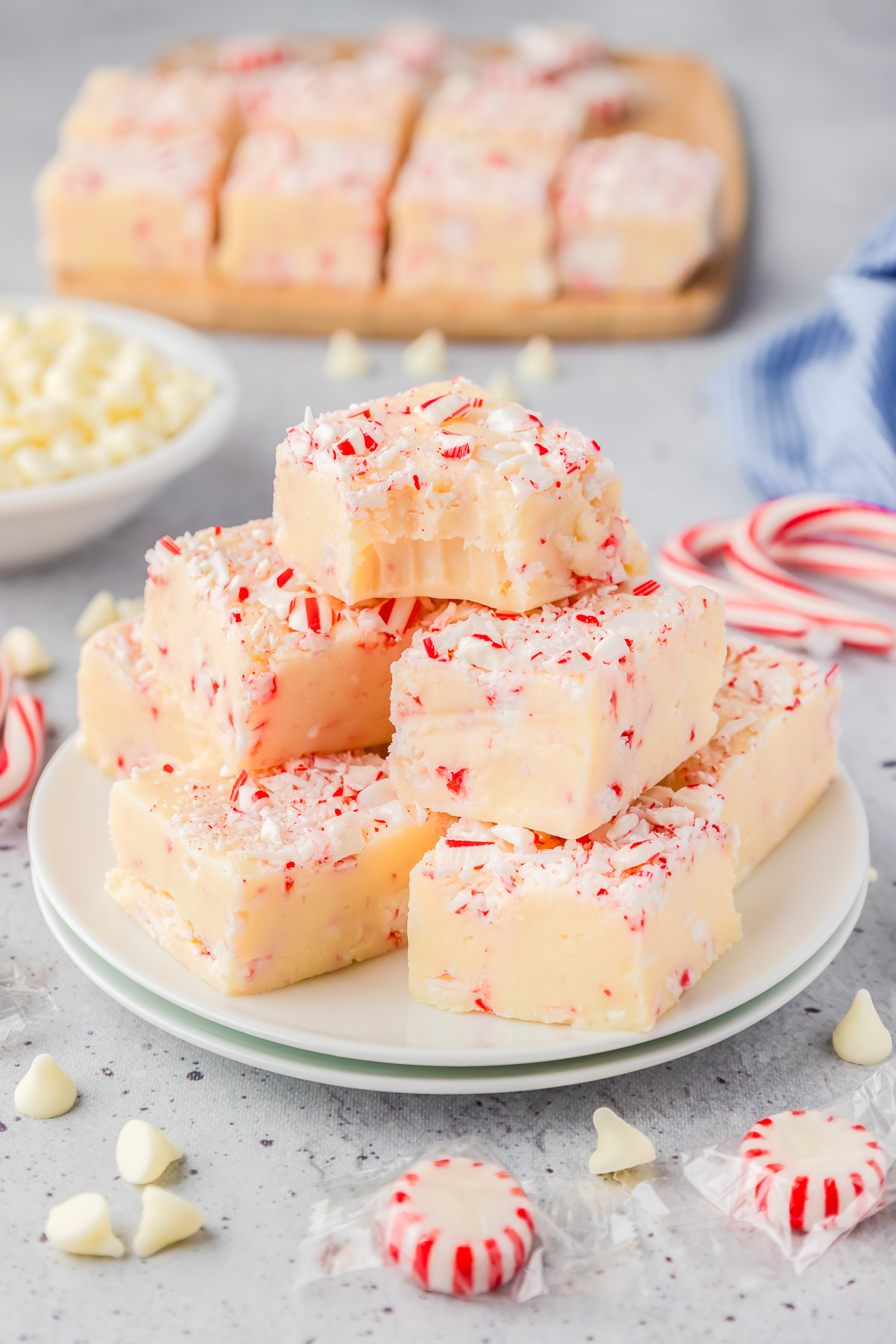Peppermint fudge topped with candy cane pieces piled on a plate with the top piece missing a bite and more candy cane fudge, candy canes, peppermint pieces and white chocolate chips on the counter.