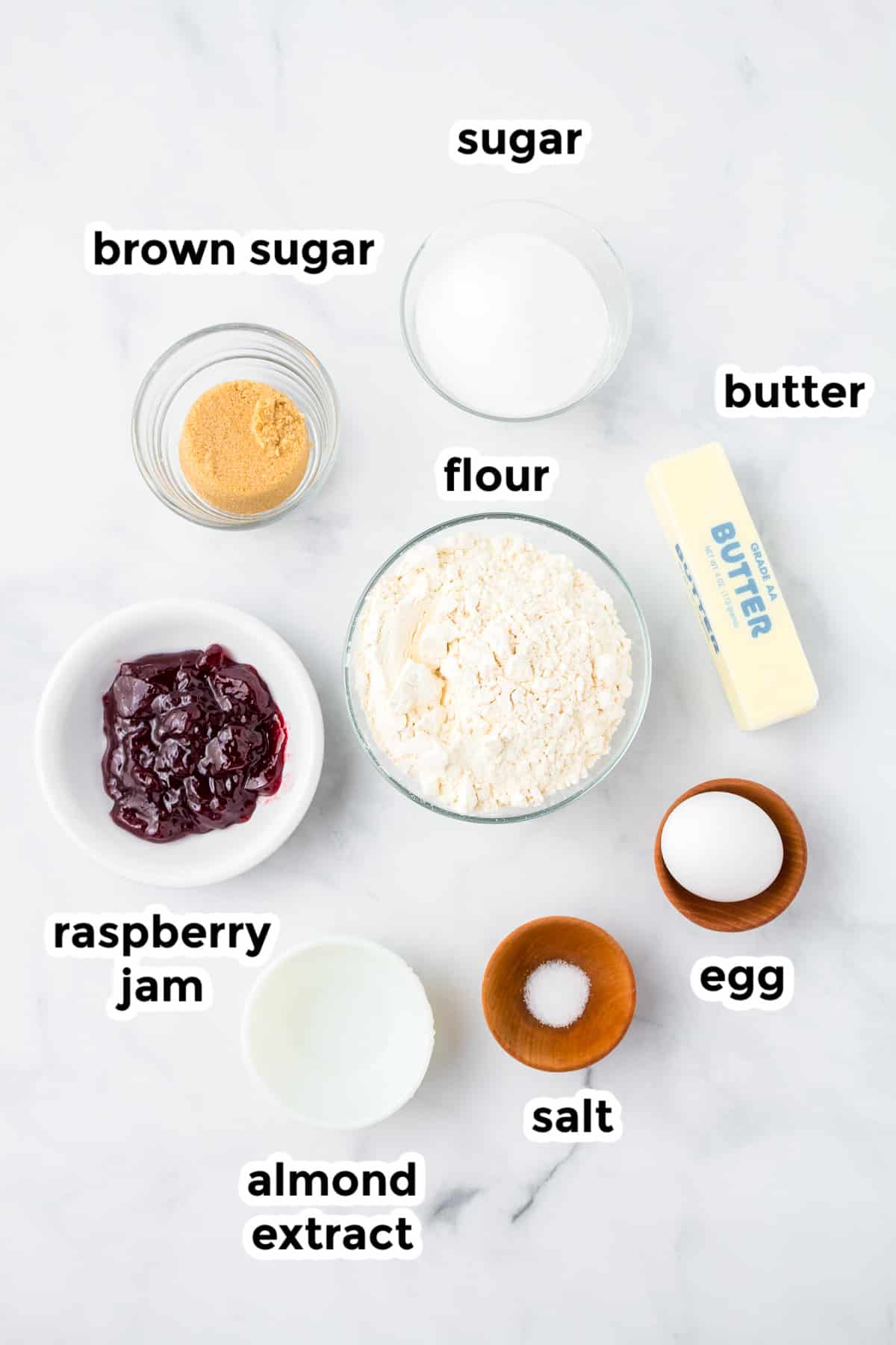 Ingredients for raspberry thumbprint cookies in bowls with text labels on a counter.