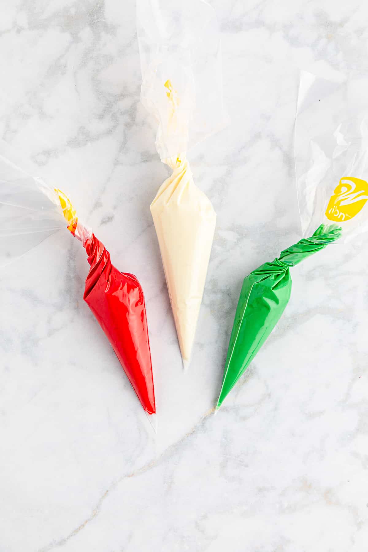 Three pastry bags full of melted red, green, and white candy melts on a marble counter from above.