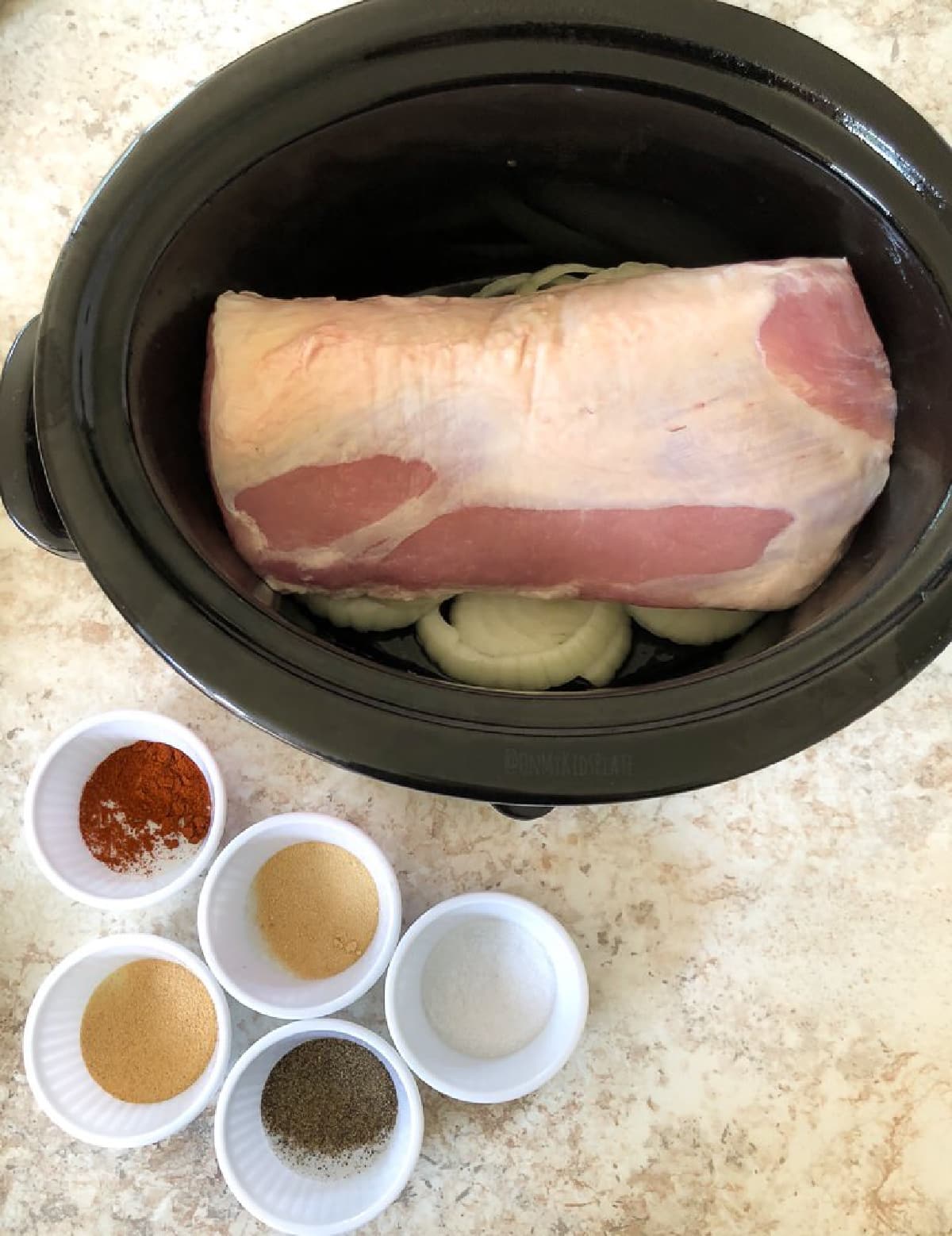 A crock pot with a piece of pork loin fat side up on top of sliced onions and five small bowls of spices on the counter nearby.