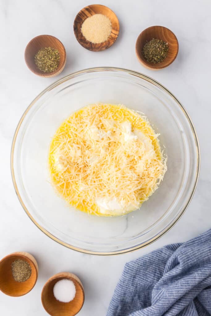 A large glass bowl filled with ricotta cheese, parmesan cheese and egg for the stuffed shell filling with small bowls of spices nearby.