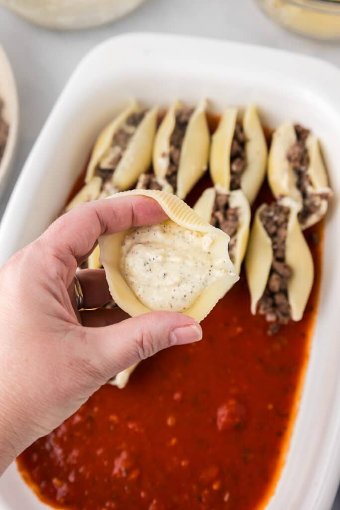 A hand holding a half full stuffed shell full of ricotta cheese that they just filled over a casserole dish.