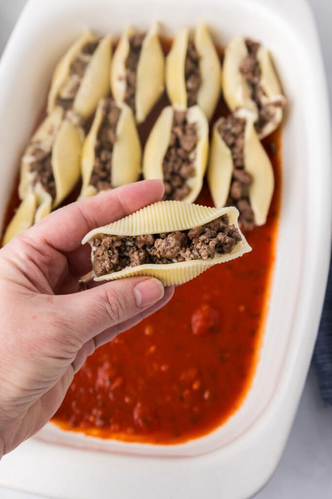 A hand holding a full stuffed shell full of ricotta cheese and beef that they just filled over a casserole dish.