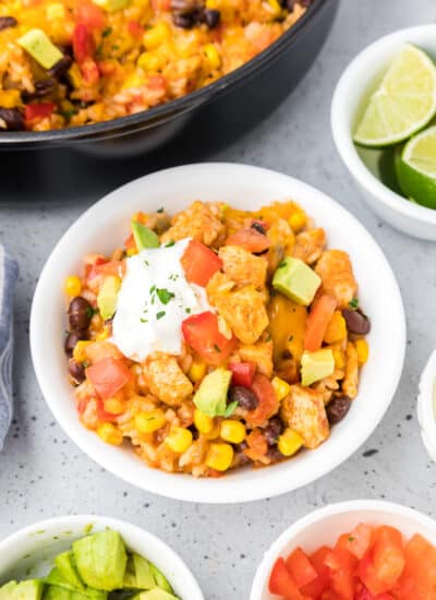 A bowl of mexican chicken and rice in a bowl with avocado and sour cream on top with more bowls of toppings and a skillet full of more rice nearby..