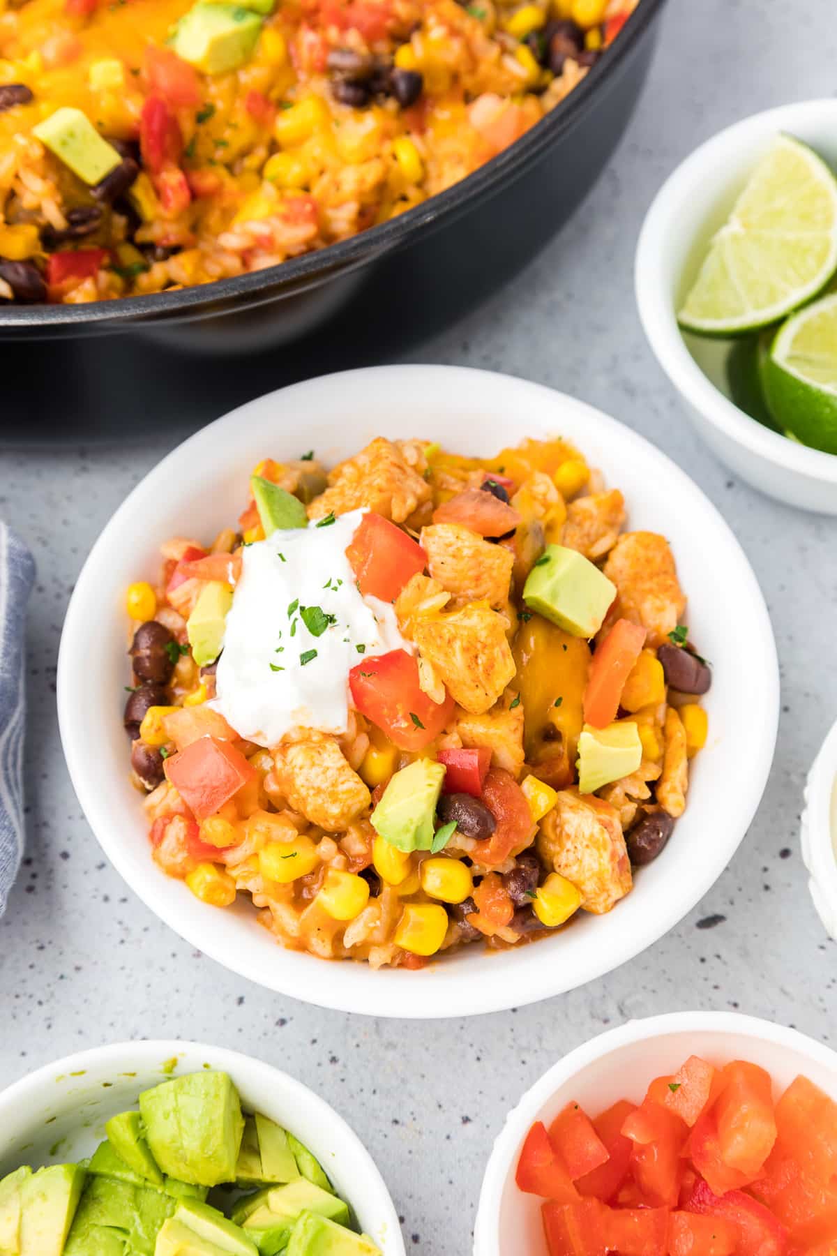 A bowl of mexican chicken and rice in a bowl with avocado and sour cream on top with more bowls of toppings and a skillet full of more rice nearby..