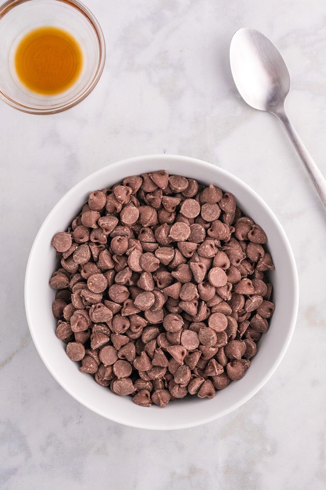 Chocolate chips in a bowl next to a small bowl of vanilla extract and a spoon.