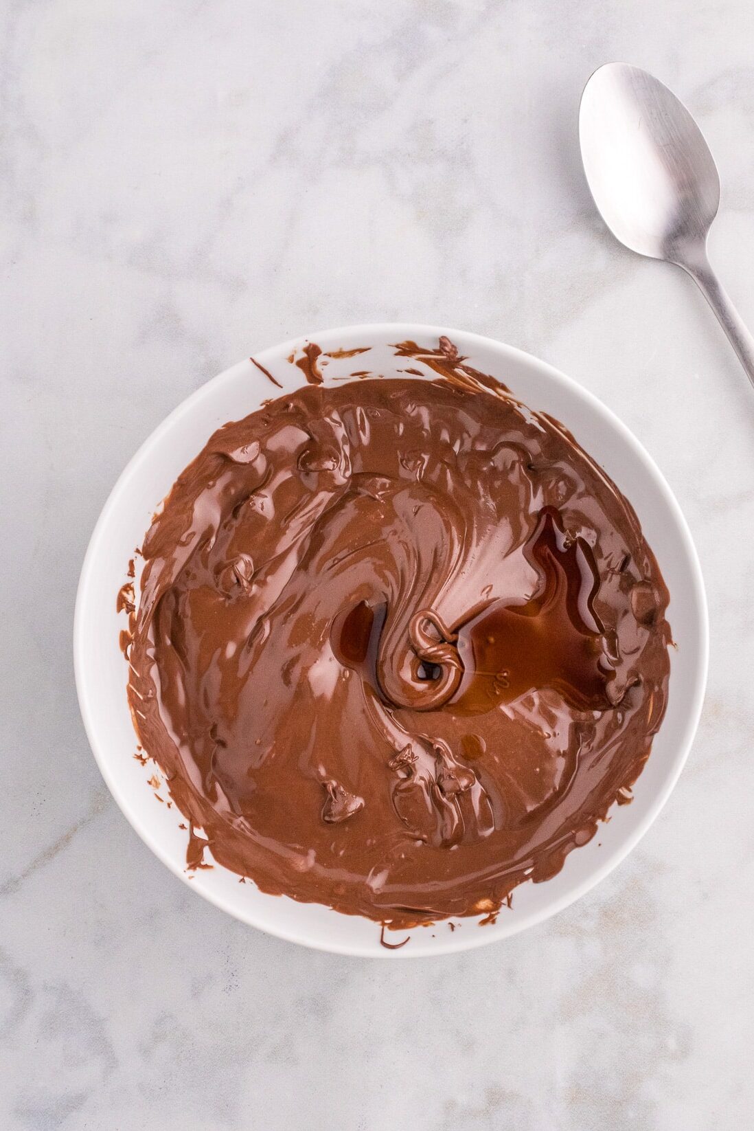 A bowl of melted chocolate sauce with a spoon next to it before stirring.
