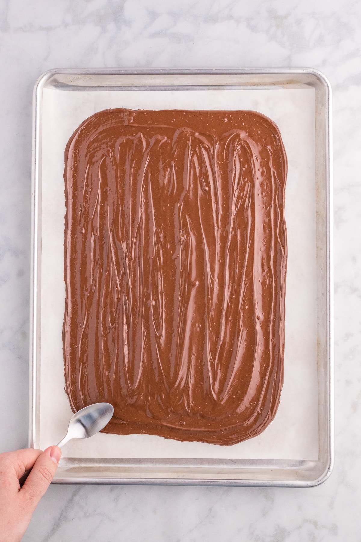 A hand spreading melted chocolate on a baking sheet with a spoon.