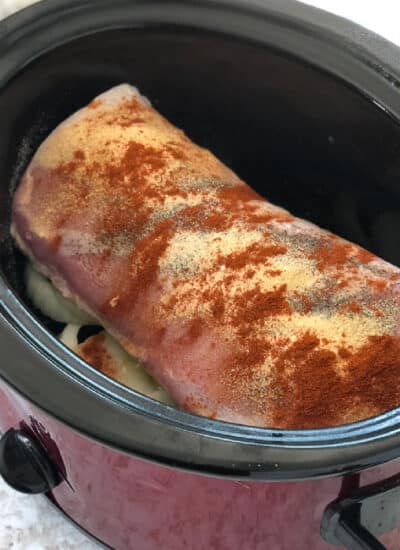 A square view of a crock pot with a piece of pork loin sitting on sliced onion and covered with spices before cooking.