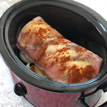 A square view of a crock pot with a piece of pork loin sitting on sliced onion and covered with spices before cooking.
