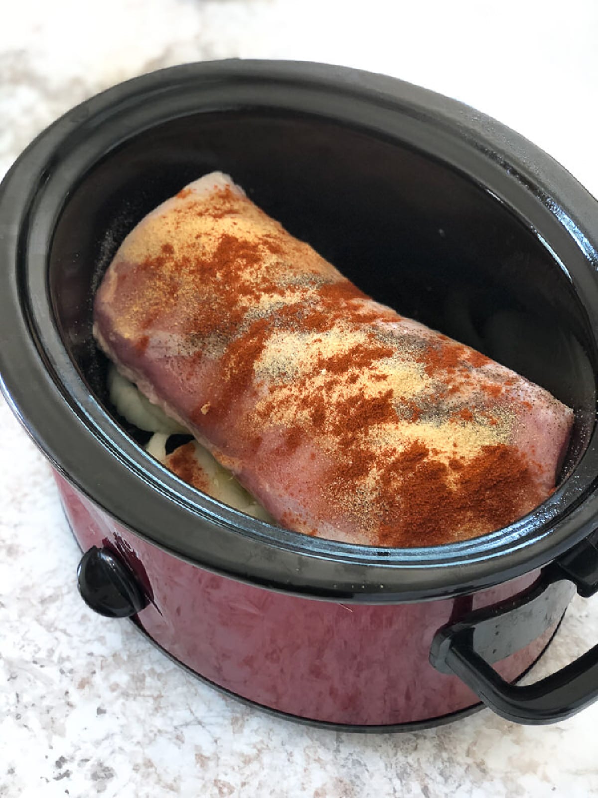 A crock pot with a piece of pork loin sitting on sliced onion and covered with spices before cooking.