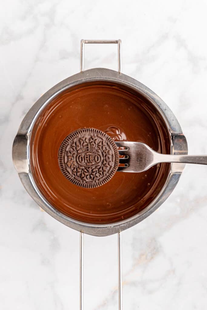 An oreo cookie being held with a fork over a bowl of chocolate.