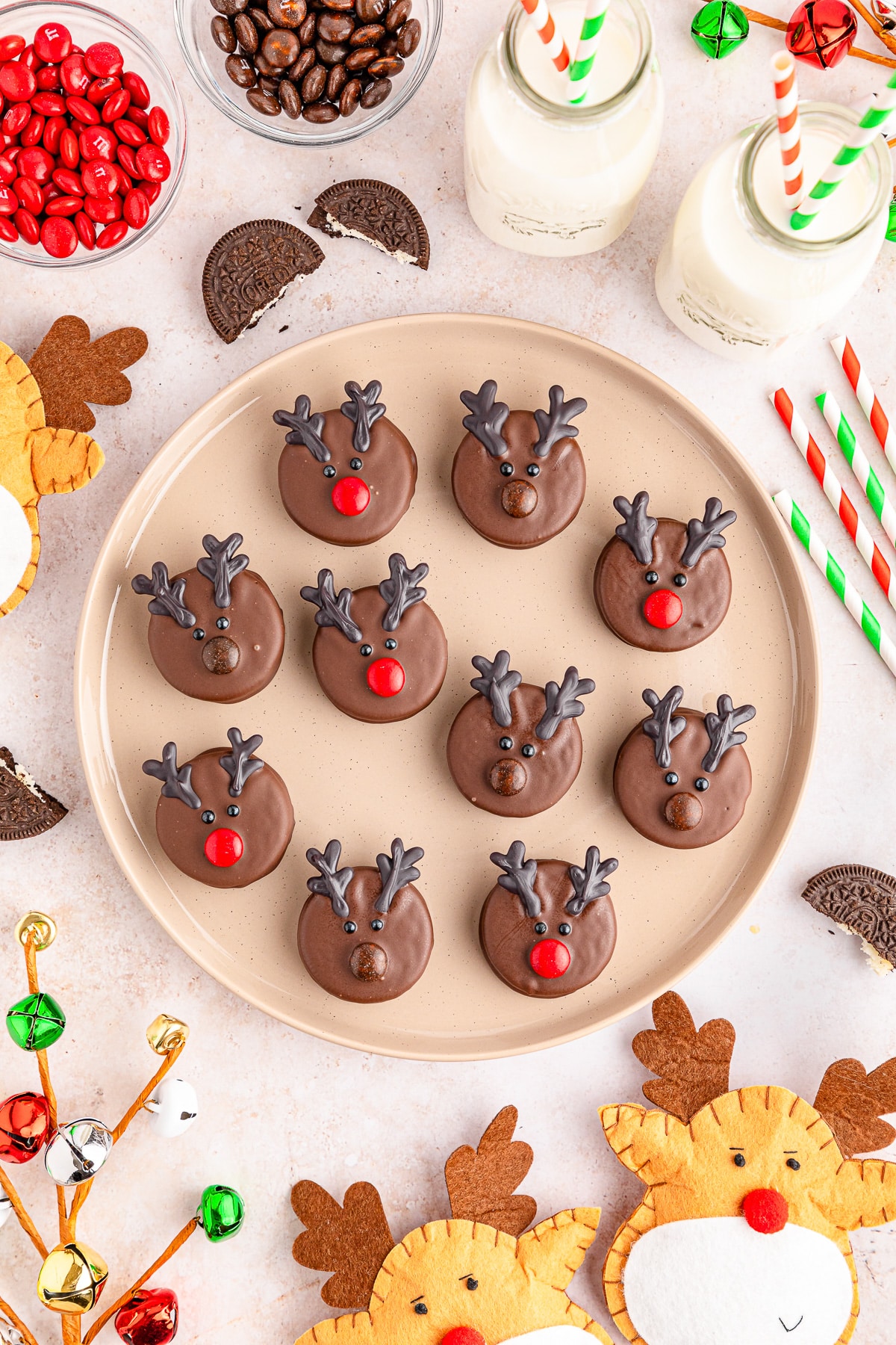 Chocolate reindeer oreo cookies on a plate with more holiday decorations and milk nearby.