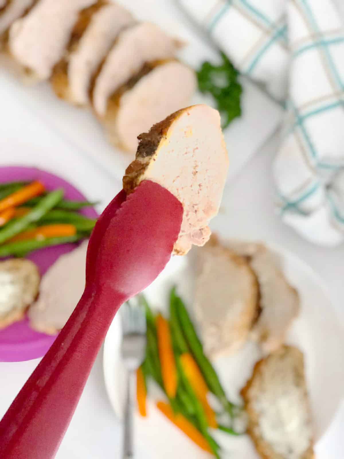 A pair of tongs holding up a slice of pork loin over top of a plater and plates full of more pork loin and green beans on the table in the background.