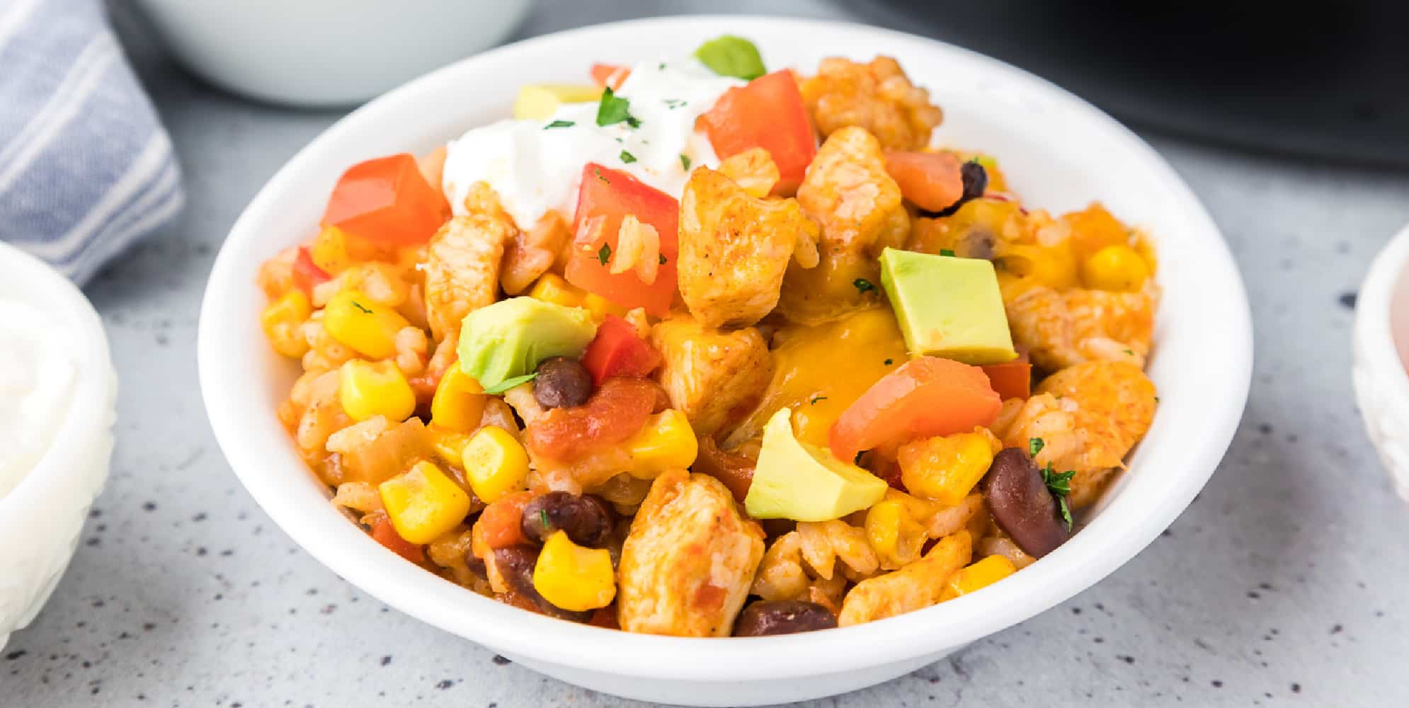 A wide view of a bowl of Mexican chicken and rice with cheese, corn, black beans, bell pepper pieces, sour cream and avocado.