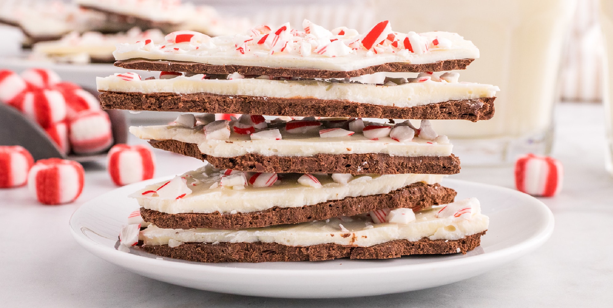 A wide view of a stack of chocolate and peppermint bark on a plat with more peppermint candies in the background.