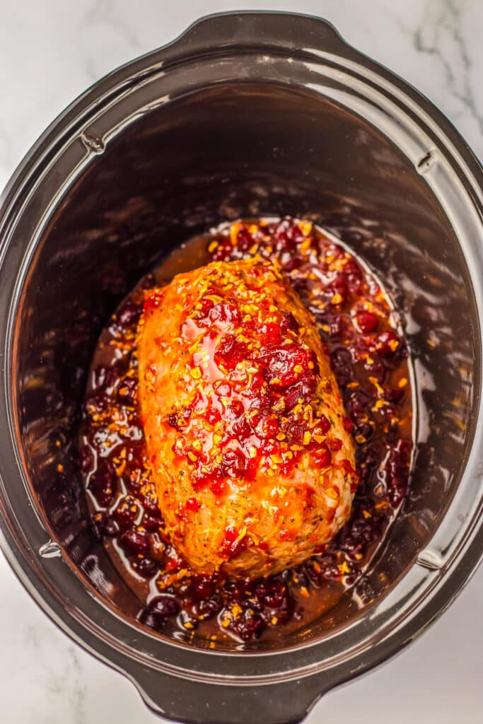A raw turkey breast in a slow cooker covered with a red cranberry orange sauce before cooking.