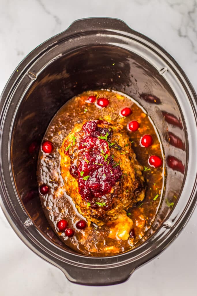 A crock pot filled with a large turkeybreast covered in cranberry sauce and cooking liquid after cooking..