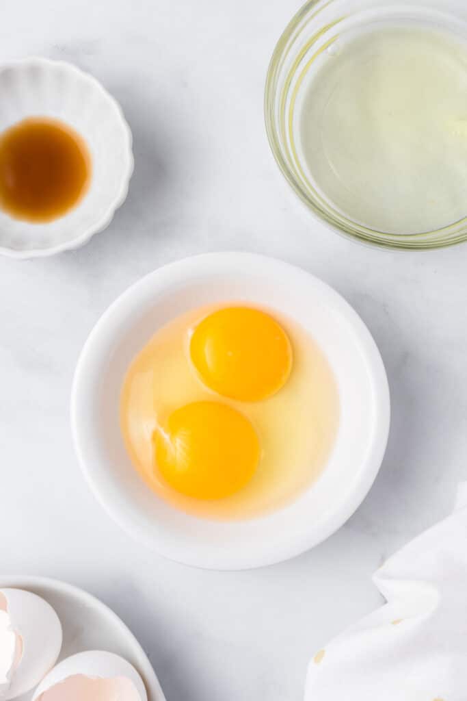 One egg with an extra egg yolk in a bowl with an egg white in a second bowl and vanilla in a third bowl nearby.