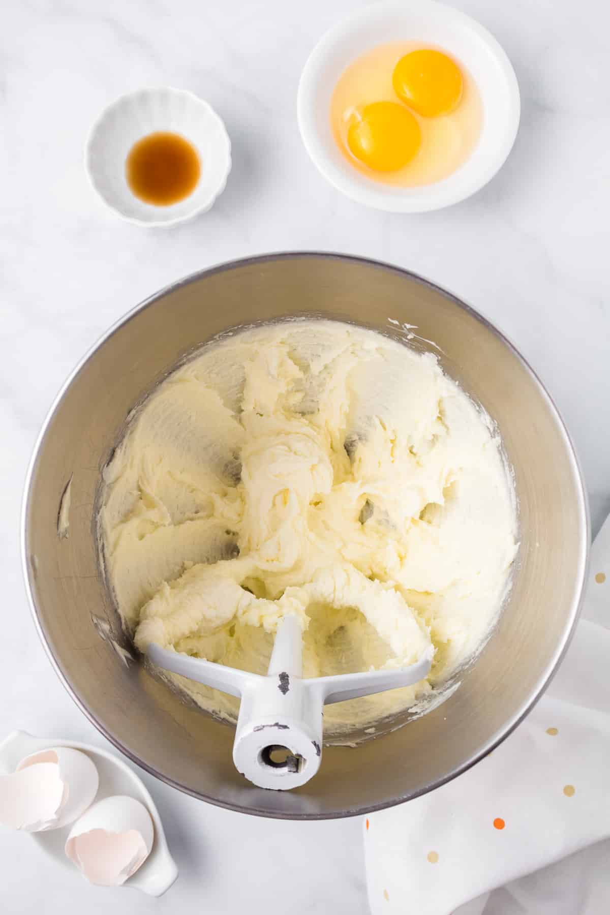 A stand mixer bowl with creamed butter and sugar in the bowl and vanilla and cracked eggs in bowls nearby.