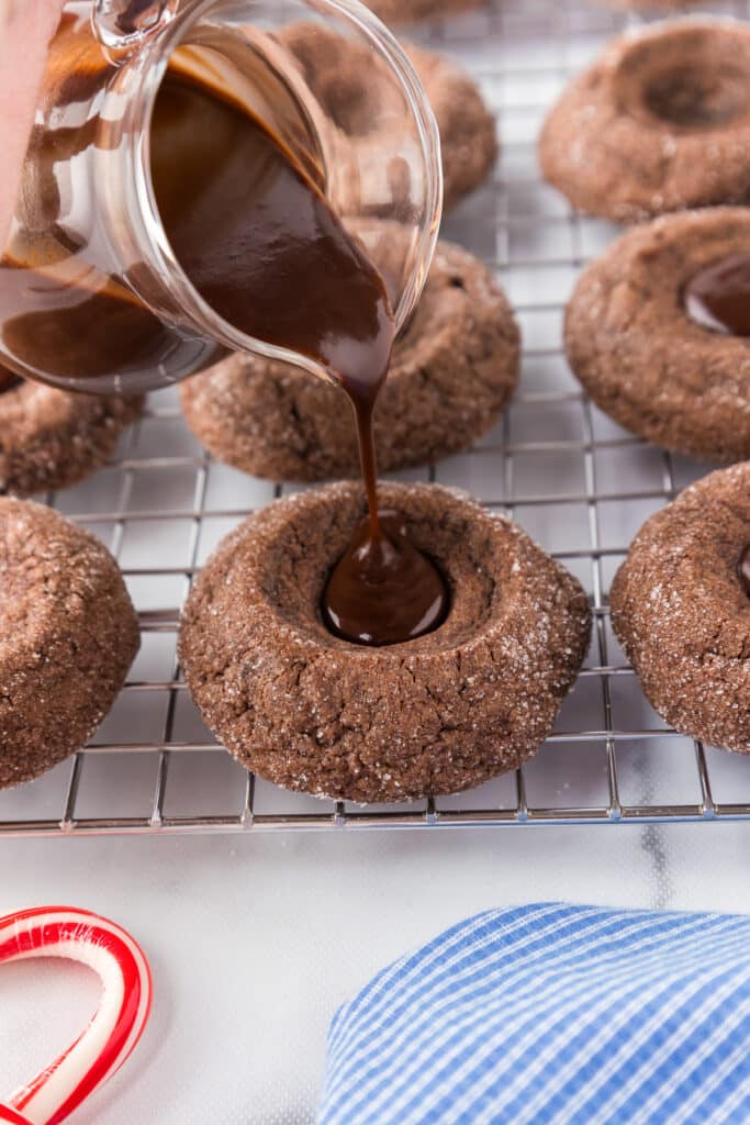 A glass measuring cup pouring liquid chocolate ganache into the center of a chocolate peppermint thumbprint cookie.
