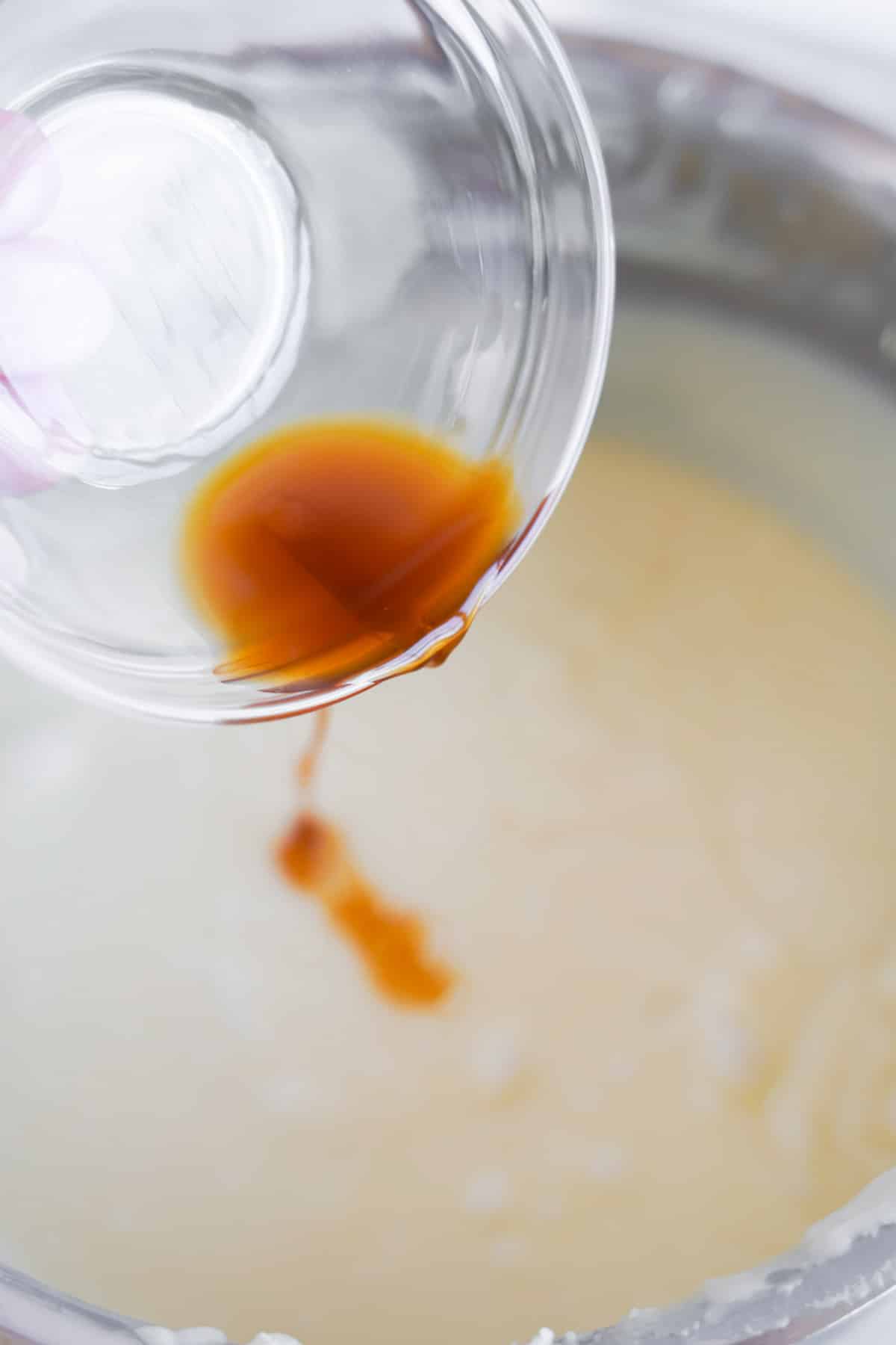 A bowl of vanilla extract being poured into a liquid vanilla fudge batter.