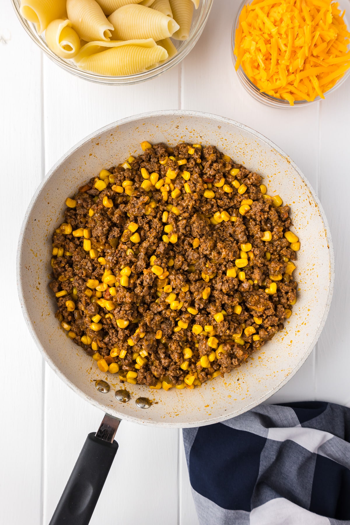A frying pan filled with ground beef and corn with cooked pasta shells and cheddar cheese on the counter nearby.