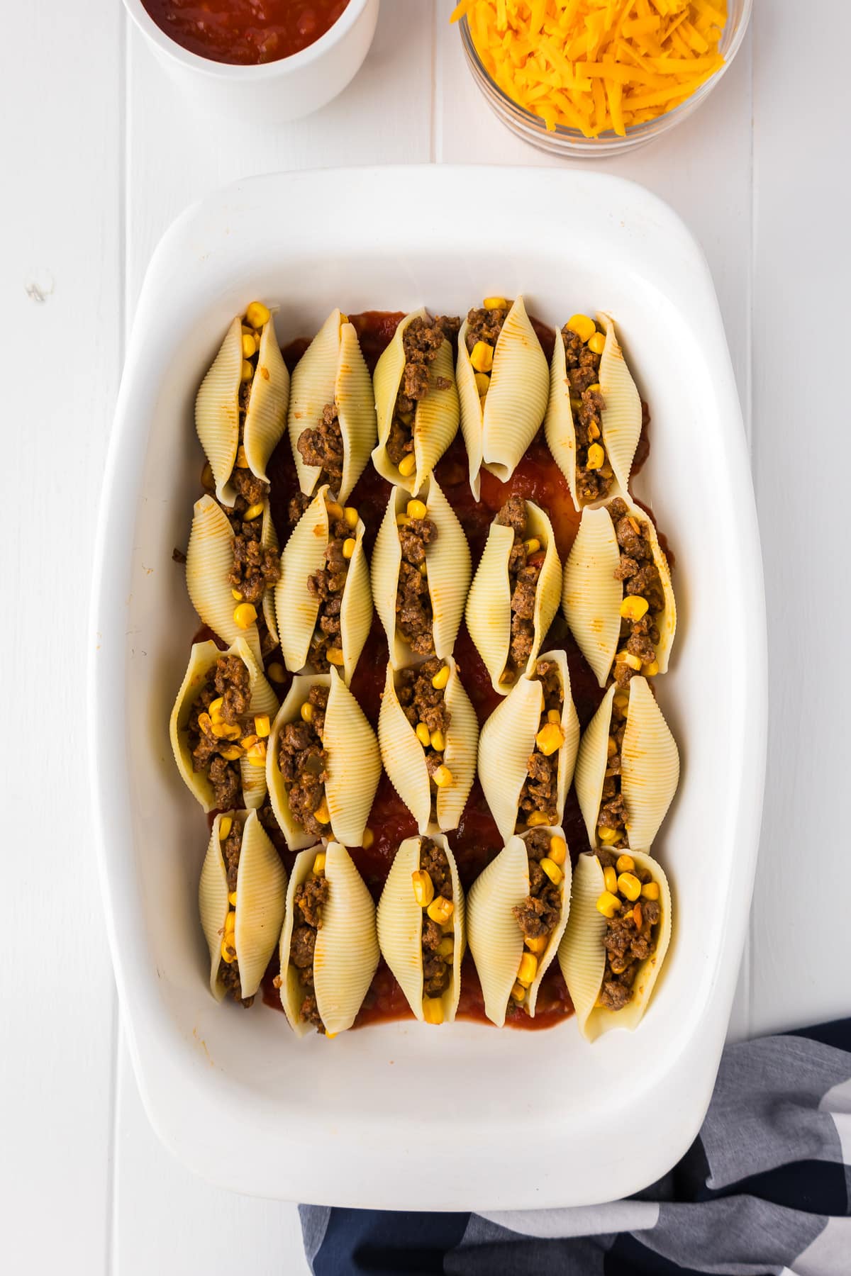 Taco shells lined up in a casserole dish stuffed with taco meat and corn with cheese and salsa in bowls nearby.