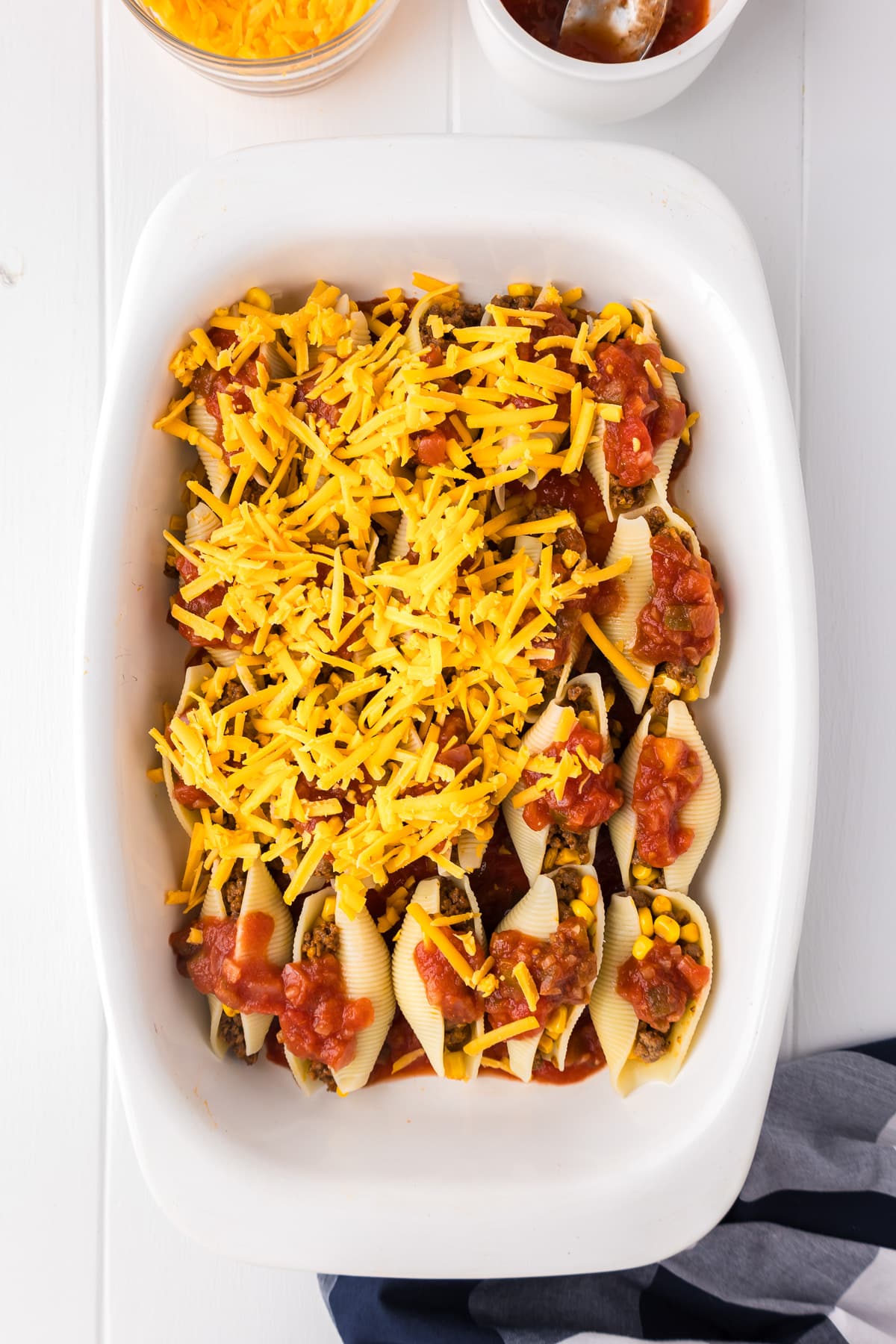 Taco stuffed shells being covered with shredded cheddar cheese.